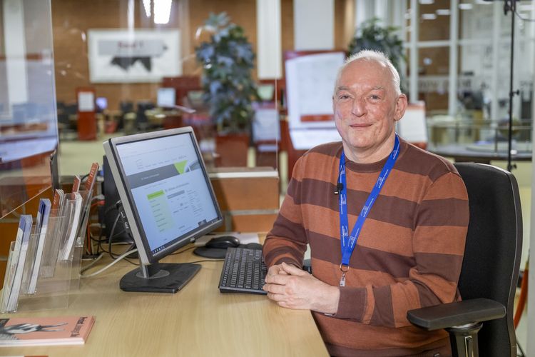 Das Foto zeigt Jens Haake. Er sitzt am Eingang der Bibliothek an seinem Computer und lächelt in die Kamera. Um seinen Hals hängt ein Schlüsselband mit dem Logo der Universität.
