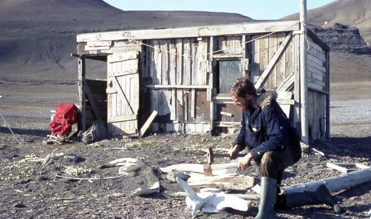 Arctic explorer Paul W.J. de Groot kneels in front of a woodshed on Edgeøya and chops wood using a whale vertebra as a chopping block. A red rucksack leans against the left side of the shed, bare mountain slopes of the island can be seen in the background. 