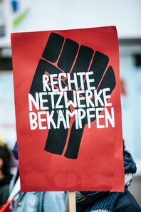 Auf dem Bild ist ein rotes Plakat zu sehen, auf dem eine schwarze Faust in die Höhe gereckt wird und in weißer Schrift "Rechte Netzwerke bekämpfen" steht.