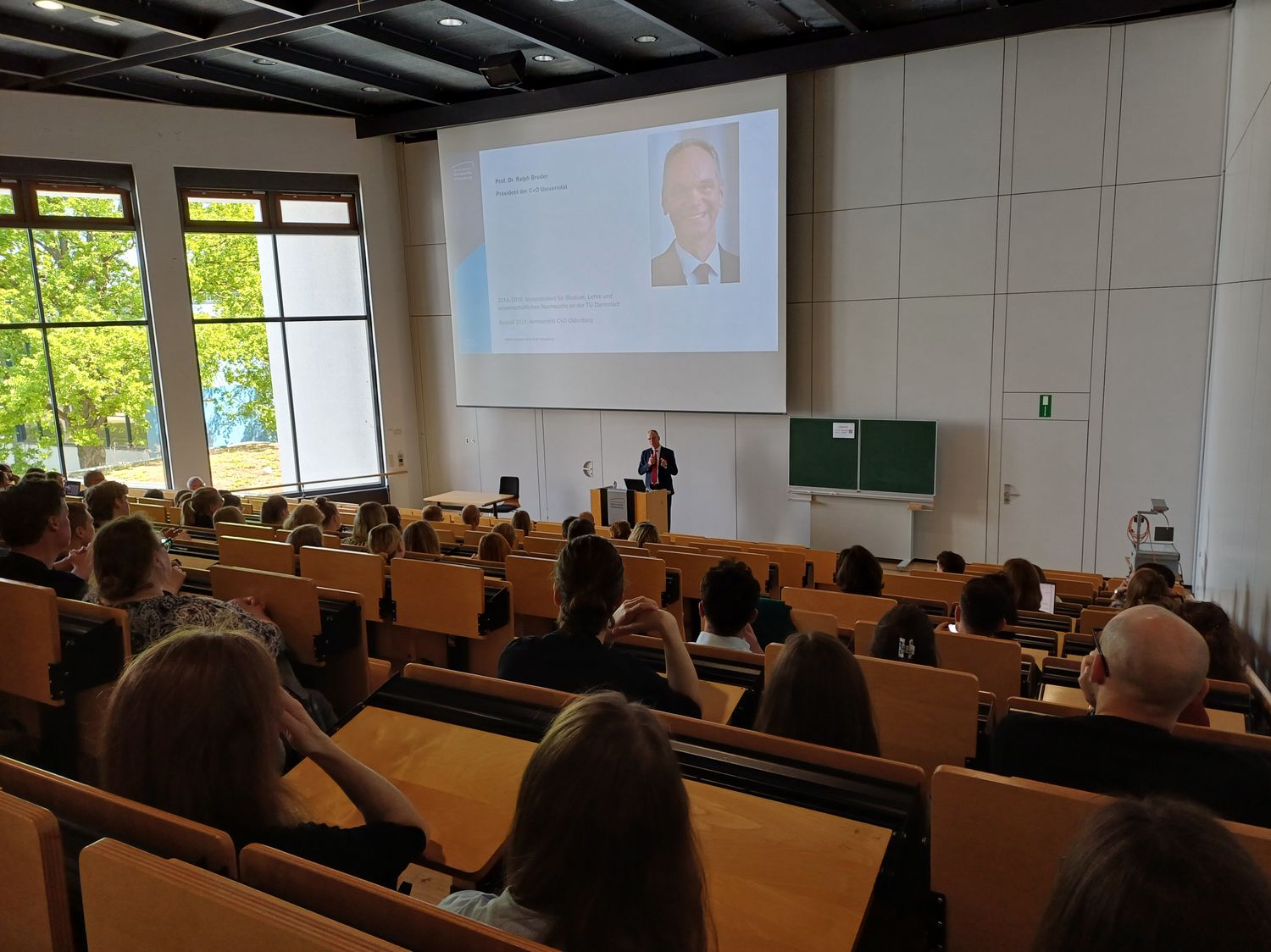 Es ist ein Hörsaal zu sehen, in dem viele Menschen sitzen. Vorne steht Universitätspräsident Prof. Dr. Ralph Bruder und hält eine Rede.