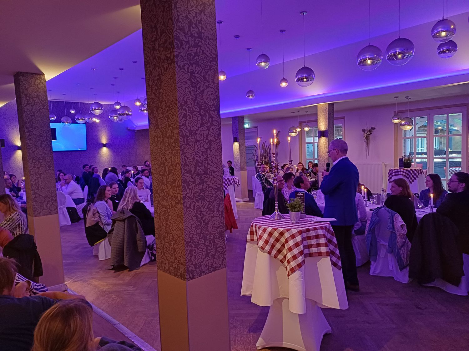 Es ist ein Festsaal zu sehen, in dem viele Menschen an runden Tischen sitzen.