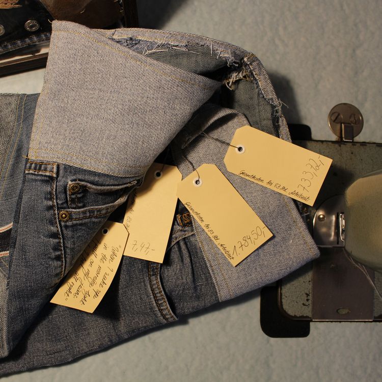 Das Bild zeigt eine Jeans, die mit verschiedenen Preisschildern ausgestattet ist. Eine Beschriftung lautet: "Gesamtkosen bei 83 Stunden Arbeitszeit: 1784 Euro und 50 Cent. 