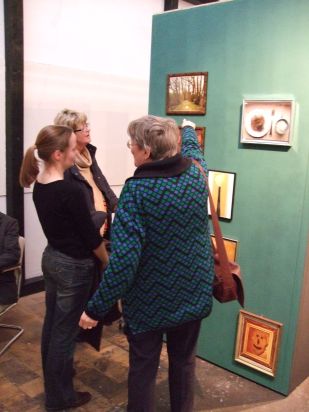 Foto der Ausstellung: Drei Menschen stehen vor einer dunkelgrün gestrichenen Wand, an der viele kleine, eingerahmte Bilder hängen.