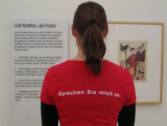 Projektfoto: Projektteilehmer*in steht vor einer Ausstellungswand. Auf dem T-shirtrücken steht: "Sprechen Sie mich an"