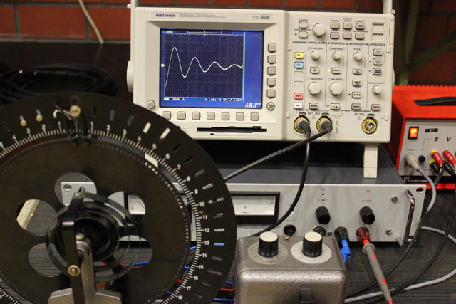 Foto eines Pohlschen Rades mit Oszilloskopbild der gedämpften Schwingungsamplitude.