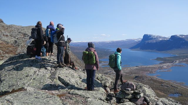The photo shows several participants of the excursion. They are standing on a mountain and looking into the distance. There are lakes and several hilltops.
