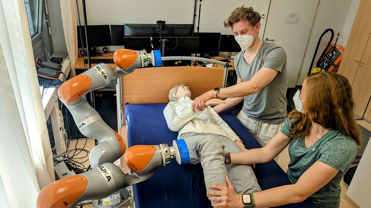 Zwei Personen stehen an einem Krankenbett und lagern eine Dummy-Puppe mit Unterstützung zweier neben dem Bett montierten Roboterarme.