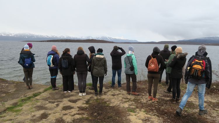 The photo shows several participants of the excursion. They are standing on the shore of a lake and looking into the distance. Several mountains can be seen on the misty horizon.
