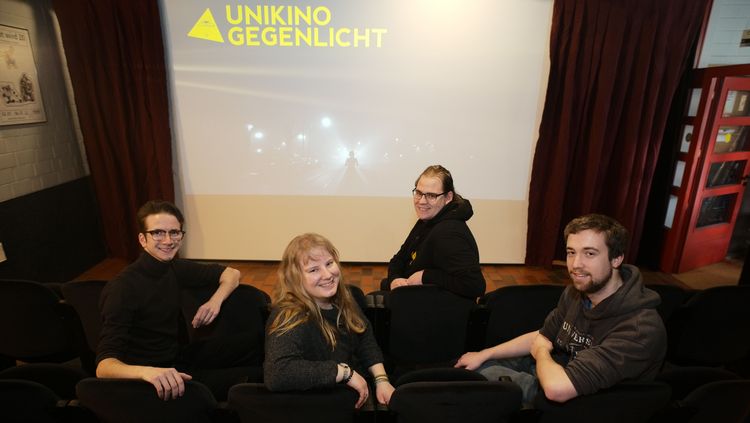 Mitglieder des Teams vom Unikino Gegenlicht sitzen auf Kinosesseln. Im Hintergrund ist die Leinwand mit dem Logo des Kinos zu erkennen.