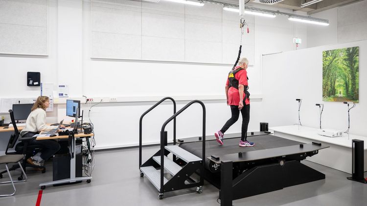 A woman is running on a wide treadmill. She is secured with a belt that is attached to the ceiling. Behind her, a young woman is sitting at a computer.