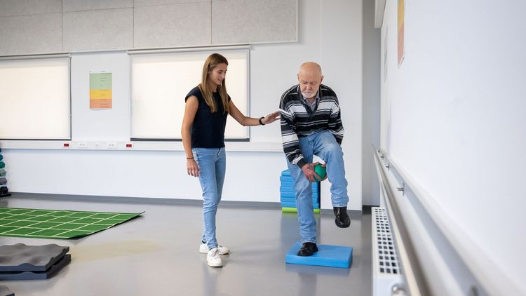 A woman assists an older man who stands on a soft mat, lifts one leg and passes a ball underneath.