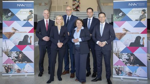 Six people in blue suits are standing in front of a staircase, with two display stands showing scenes from the Northwest Alliance's research standing beside them.