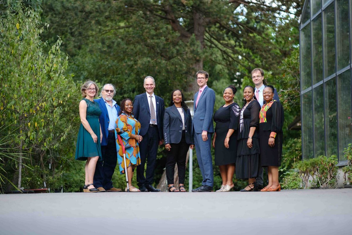 Das Bild zeigt eine Gruppe von zehn Personen, die für ein formelles Foto posieren. Die Gruppe besteht aus Vertreter*innen der Universität Oldenburg und der Nelson Mandela University im Rahmen des 25-jährigen Kooperationsjubiläums. Sie stehen vor einem Hintergrund aus Grün und einem Gewächshaus. Die Personen sind festlich gekleidet und vermitteln einen Eindruck von Würde und Zusammenarbeit.