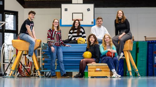 Sieben junge Menschen sind in einer Turnhalle. Sie sitzen auf Mattenwagen und Sprungbock.