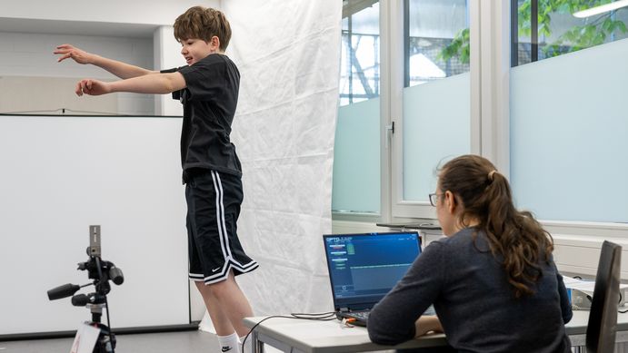 Kind springt in die Luft. Erwachsene Person sitzt hinter Laptop und beobachtet Jungen.