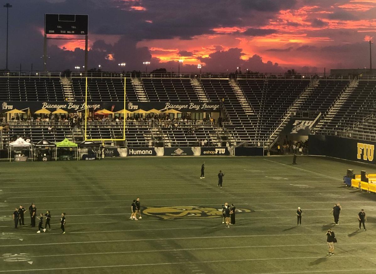Footballstadion bei Sonnenuntergang, wenige Menschen auf dem Spielfeld und Tribünen.