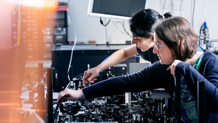 Zwei Frauen bereiten in einem Physiklabor ein Experiment vor, in dem sie mikroskopähnliche Geräte einrichten..