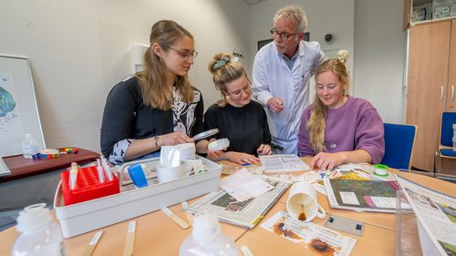 The three students are sitting at the table, which is full of medical materials such as mouth spatulas, syringes and infusion containers. Behind them stands a man in scrubs.