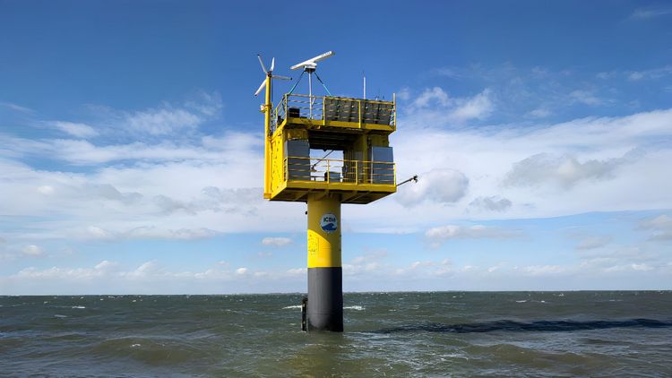 Auf dem Bild ist der Messpfahl im Meer zu sehen, der Himmel ist blau mit ein paar Wolken. An der Spitze des Pfahls befindet sich der COntainer. Er hat in der Mitte einen offenen Bereich, auf dem Dach befinden sich einige Messgeräte.