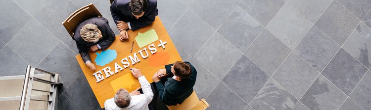 Eine Draufsicht von vier Personen, die an einem Tisch mit dem Schriftzug „ERASMUS+“ darauf sitzen.