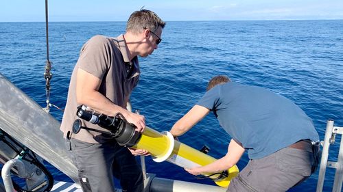 Two men stand at the stern of a ship and throw the yellow, elongated buoy into the sea.