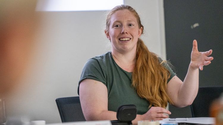 Mira Richts has long reddish-blonde hair tied back in a ponytail that falls over her shoulder. She is wearing a green T-shirt. As she speaks, she gestures with her left hand.