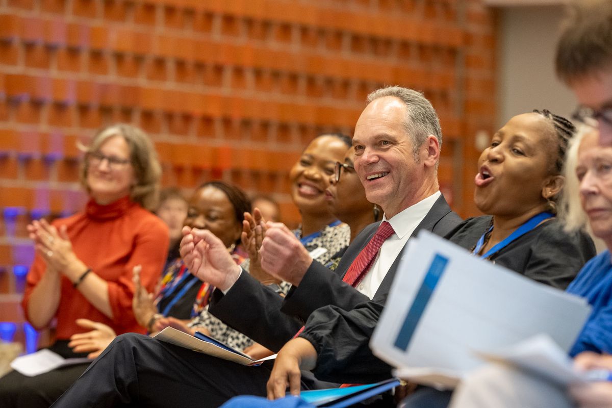 Das Bild zeigt eine Gruppe von Menschen, die im BIS-Saal sitzen und klatschen. Im Vordergrund sitzt ein Mann in einem Anzug mit einem freundlichen Lächeln und klatscht mit den Händen. Neben ihm sitzen mehrere Frauen, die ebenfalls lächeln und klatschen. Im Hintergrund sind weitere Personen zu sehen. 