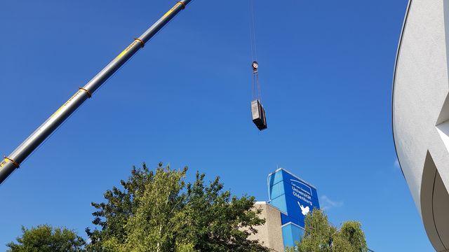 View into the sky, in the foreground a computer cabinet hangs from a crane, in the background trees and the A1 building with a dove of peace, on the right the round wall of the lecture theatre centre still protrudes into the picture. 