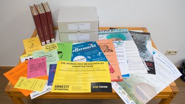 Several colourful leaflets, posters and brochures lie on top of each other on a table, with three volumes and two boxes from the leaflet collection at the back.