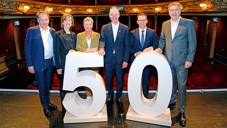 Sechs Personen stehen hinter einer großen 50 auf der Bühne des Staatstheaters, im Hintergrund der leere Publikumsbereich des Theaters. 