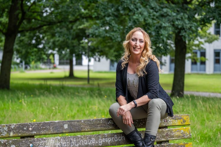 Julia Michaelis sitzt auf der Lehne einer Bank. Sie beugt sich leicht nach vorne und lächelt in die Kamera. Im Hintergrund sind das Gebäude A 5 sowie eine Rasenfläche und einige Bäume zu erkennen.