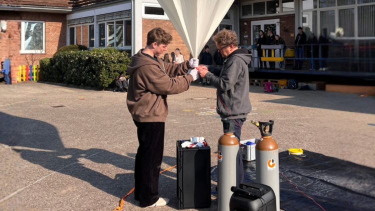 Junger Mann in brauner Jacke links und mittelalter Mann in grauer Jacke rechts halten gemeinsam einen Wetterballon fest.