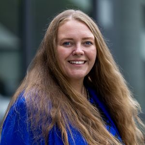 Porträtfoto von Frau mit langem dunkelblondem Haar und königsblauem Jackett lächelt in Kamera. 