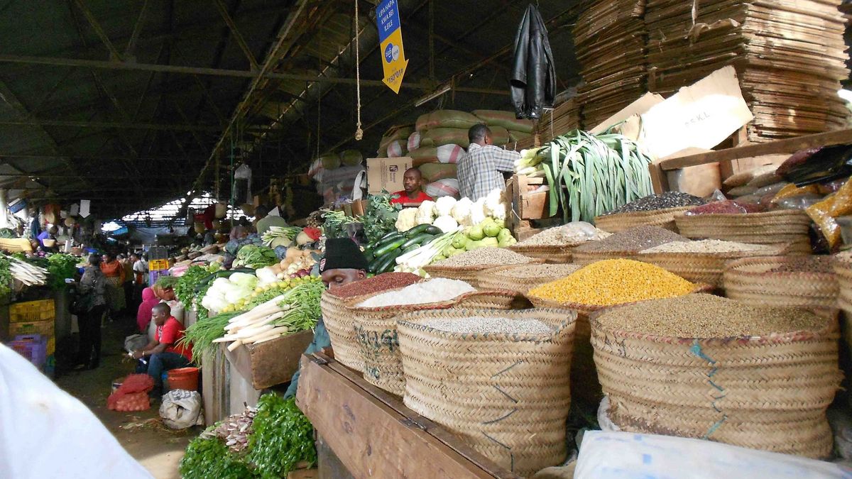  Ein belebter Marktstand in Dar es Salaam mit frischen Produkten wie Hülsenfrüchten, Getreide und Gemüse. Menschen stehen und gehen zwischen den Ständen.