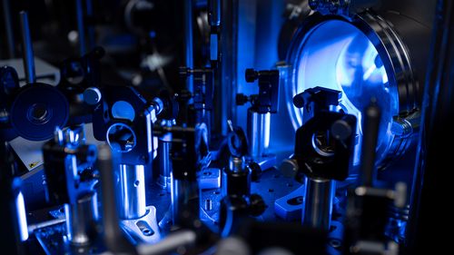 The picture shows a close-up of the experimental setup. On the right is the vacuum chamber, a stainless-steel tube with a glass window. In front of it are various optical instruments, such as prisms and lenses, which are used to direct the two laser beams.