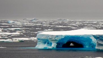 The image shows the Southern Ocean. The sea and sky are grey and there are lots of ice floes drifting on the sea, as well as a single jagged iceberg made of blue glacier ice.