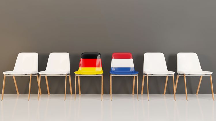 Sechs nebeneinander stehende Stühle deuten ein Wartezimmer an. Die beiden mittleren Stühle sind nicht weiß. Einer trägt das deutsche, einer das niederländische Flaggenmuster.