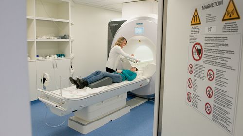 A patient is lying on a couch in front of the magnetic resonance tomograph. A technician is installing a coil above her head, which is used for the measurement.