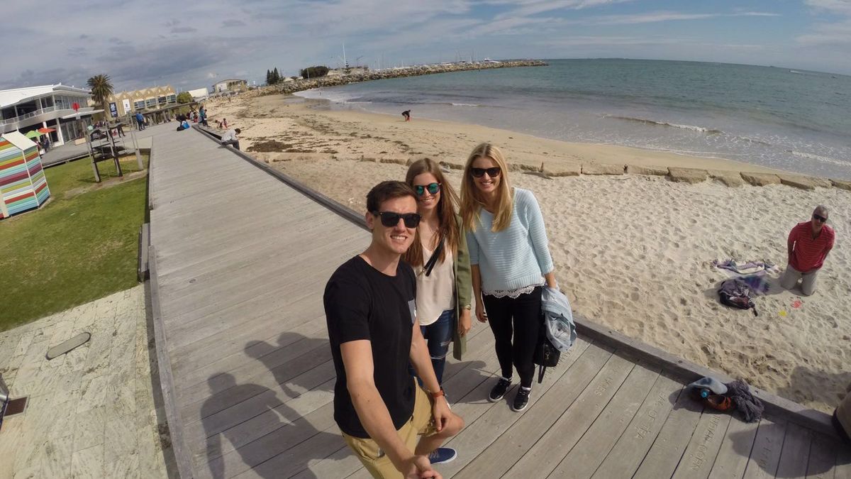 Drei lächelnde Personen posieren auf einem Holzsteg am Strand.