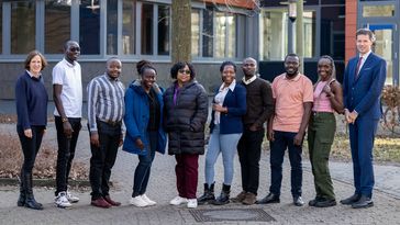 Das Bild zeigt die Gruppe der DAAD-Stipendiatinnen und -Stipendiaten. Gemeinsam mit Malve von Möllendorff und Karsten Speck stehen sie vor einem Unigebäude und lächeln in die Kamera. Insgesamt befinden sich zehn Personen auf dem Bild.