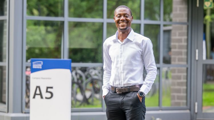 Emmanuel Asane-Otoo stands in front of the entrance to building A 5, smiling at the camera and holding his hands in his trouser pockets. In the background, bicycles are reflected in the window panes.