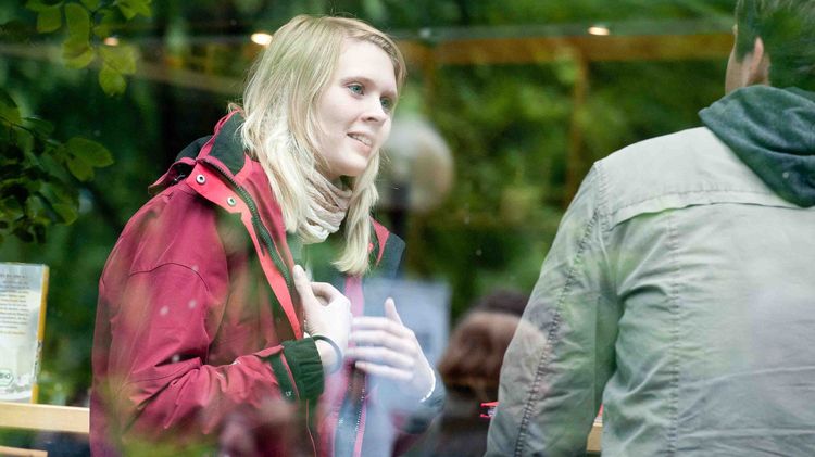 Eine Frau und ein Mann werden durch eine Glasscheibe betrachtet. Die Frau mit roter Jacke gestikuliert.