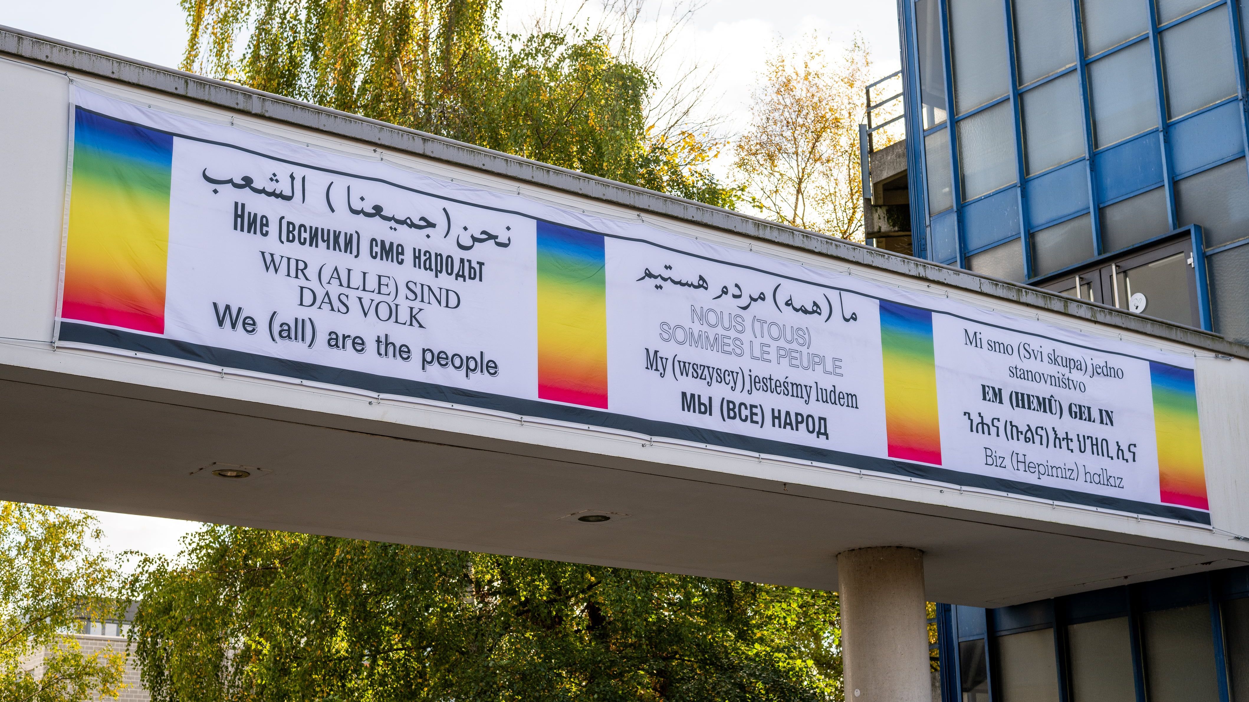 The image shows the front of the artwork "We (all) are the people". The words are written in twelve languages on a white banner hanging from the bridge between building A1 and the lecture hall centre. The rainbow colours are displayed to the left and right of the words, which are divided into three blocks.