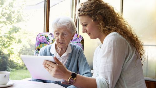 A younger woman sits at the table with an older woman and explains something to her on the tablet.