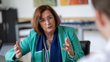 Frau Strübind sitzt dem Interviewer gegenüber. Sie sitzen im Sitzungssaal des Senats, im Hintergrund sind bunte Gemälde zu erkennen. Frau Strübind gestikuliert mit ihren Händen, während sie etwas erläutert.