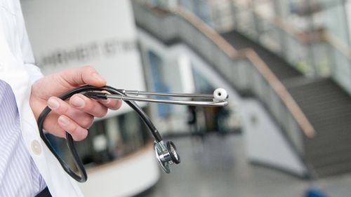 A person in a lab coat holds a stethoscope. The staircase of the University of Oldenburg's lecture theatre centre can be seen in the background.