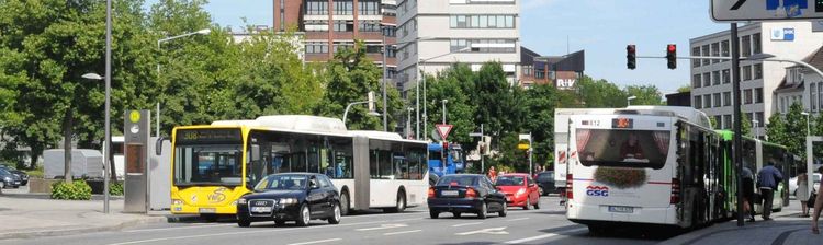 Das Bild zeigt eine Straßenszene in Oldenburg. Im Vordergrund sind mehrere Autos und Busse zu sehen, die auf der Straße fahren. Im Hintergrund erstrecken sich hohe Gebäude, darunter ein modernes Gebäude mit einer roten Fassade. Über der Straße befinden sich Verkehrsschilder, die auf Parkplätze hinweisen. Auf dem Gehweg sind Fußgänger zu sehen. 