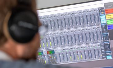 A man with headphones is sitting in front of a computer monitor that displays many controls. The viewer looks over the man's shoulder.