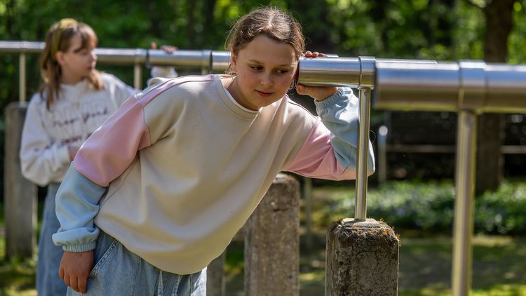Mädchen im cremefarbenen Pullover hält ihr Ohr an ein Metallrohr und lauscht.