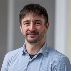 A man with a short beard and brown hair that falls slightly onto his forehead, wearing a light blue shirt, smiles at the camera.